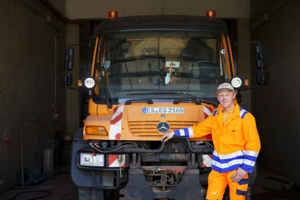 Ein Bauhof-Mitarbeiter in orangener Arbeitskleidung steht in einer Garage vor einem orangen Bauhof-Lastwagen