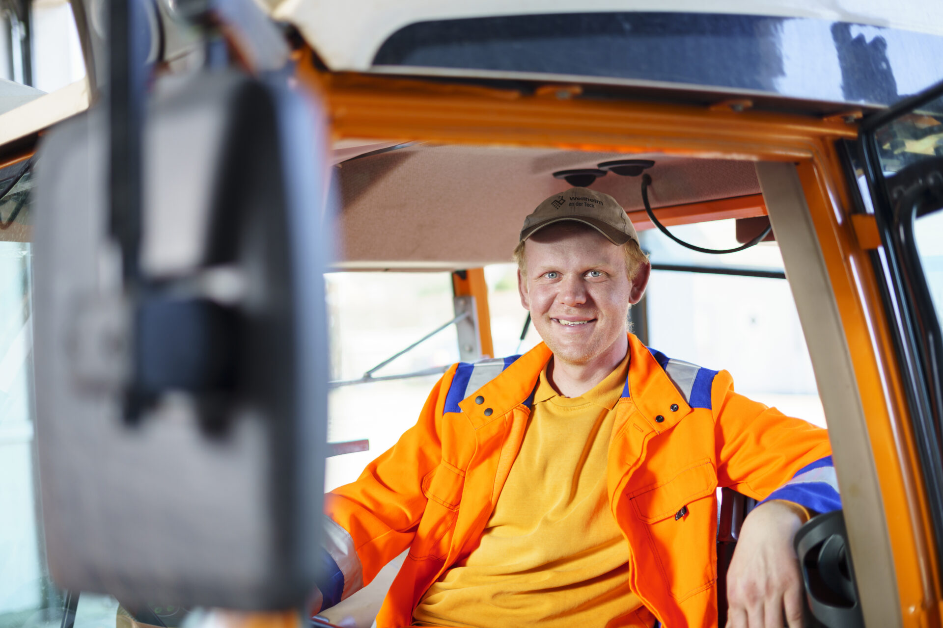 Ein Bauhof-Mitarbeiter in orangener Arbeitskleidung sitzt lächelnd auf dem Fahrersitz eines Lastwagens