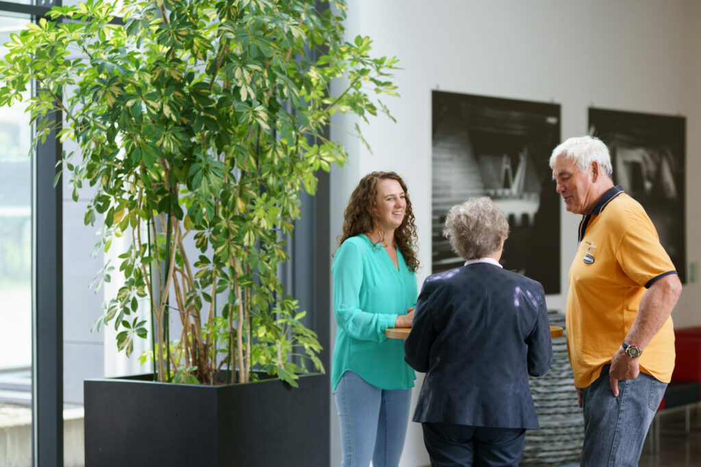 Eine Verwaltungsmitarbeiterin der Stadt Weilheim an der Teck an einem Stehtisch im Gespräch mit zwei Senioren, neben einer großen grünen Zimmerpflanze