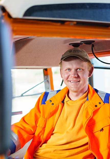 Ein Bauhof-Mitarbeiter in orangener Arbeitskleidung sitzt auf dem Fahrersitz eines Lastwagens und lacht